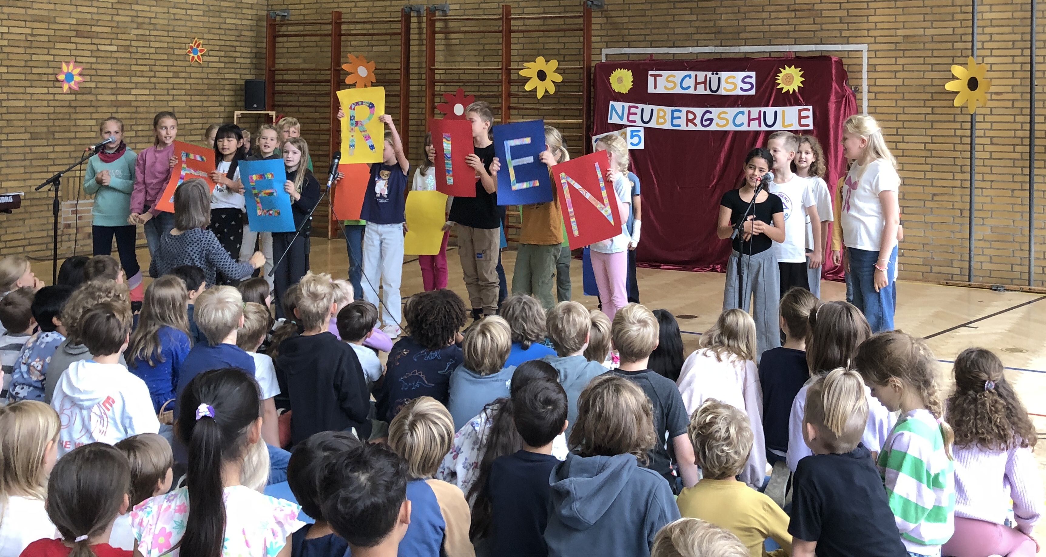 Kinder der Klasse 3a beim Performen des Ferienlieds. Sie halten bunte Schilder mit Buchstaben hoch, die das Wort „Ferien“ bilden. 